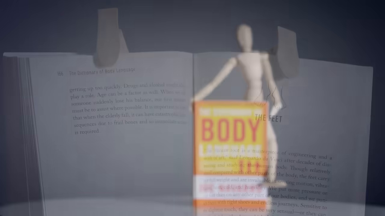 [04:19] A wooden mannequin holds up a copy of the book "The Dictionary of Body Language" by Joe Navarro. —