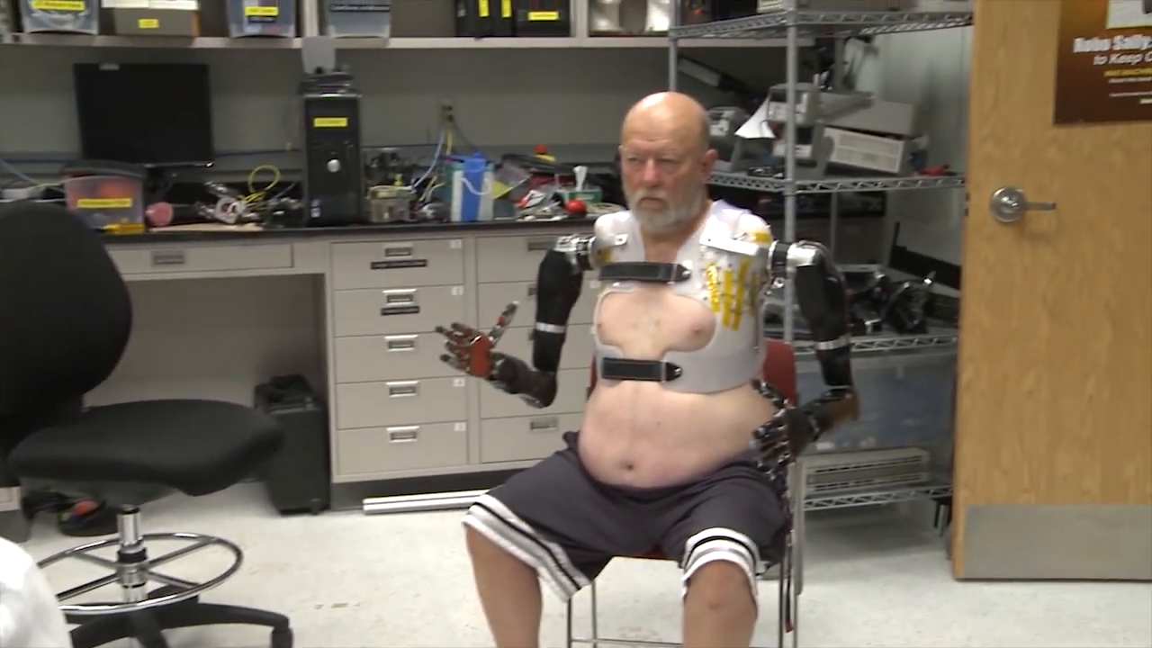 A wide shot showing the patient sitting with both advanced robotic arms fully attached to his torso harness. -