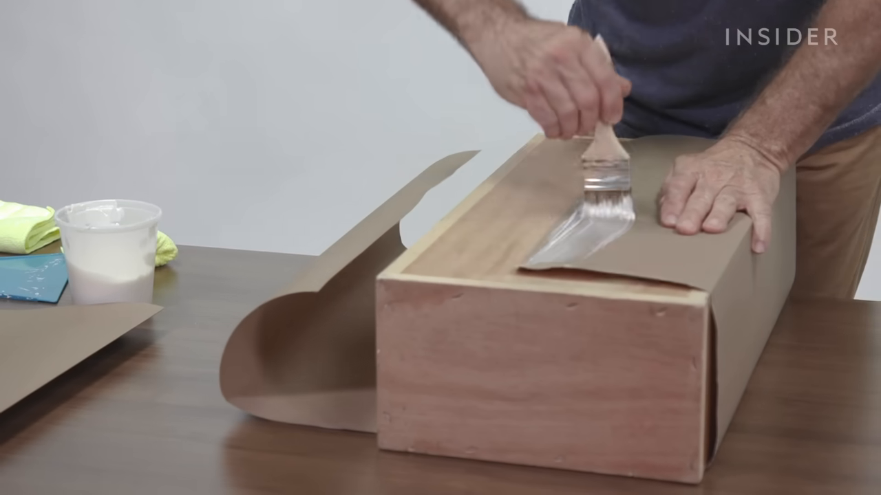 A demonstration shot of a man using a brush to apply glue to fibrous brown paper folded around a wooden mold to create a fake paper bag.